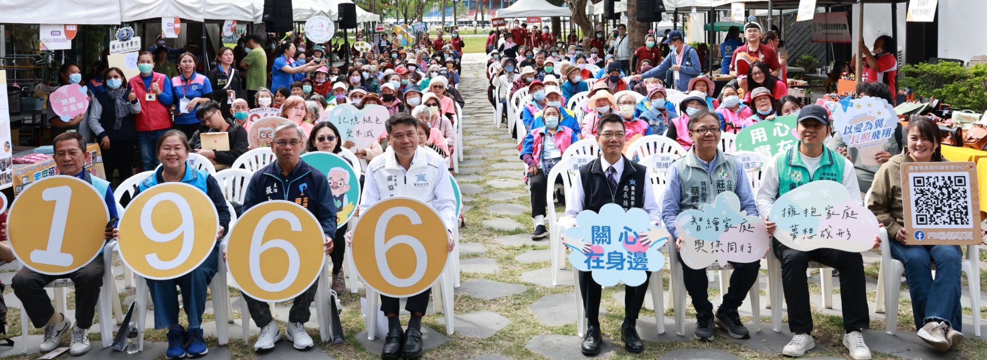 這是一張戶外活動的大合照，主要內容是一群人坐在白色塑膠椅上，面向鏡頭微笑。前排有八位穿著工作服或背心的男女，各自手持象徵意義的圓形或雲朵造型牌子，顏色有黃色、藍色、白色等。前面四人手中分別舉著數字「1966」的黃色圓牌，象徵長照服務專線。其餘四人持有標語，如「關心在身邊」、「擁抱家庭 夢想成行」等。背景有許多長者和志工穿著鮮明的藍色、桃紅色，部分人戴口罩，顯得重視健康防護。後方可見簡易棚架和各種宣傳牌，還有工作人員忙碌穿梭。