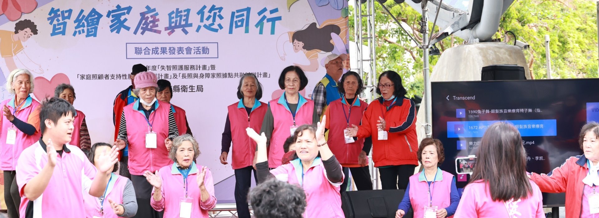 這是一張戶外活動的照片，背景是一個大型布條，上面用藍色大字寫著「智織家庭與您同行」活動名稱，並有花朵和插畫裝飾。中間站著一群穿著紅色背心和藍色領口制服的長者志工，胸前掛有識別證。前方有些人正舉手拍手，場面氣氛活潑。右側有一座大型螢幕顯示影音檔案，現場有許多綠樹環繞，光線明亮，帶來溫暖歡樂且充滿關懷的氛圍。