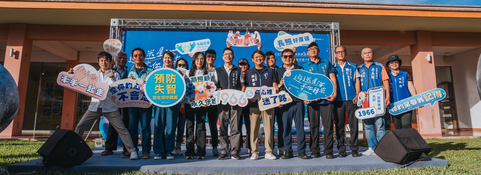 戶外活動現場合照，一群約二十位民眾與工作人員站在舞台前合影。背景為活動舞台與藍色宣導背板，上方可見晴朗藍天。多位參與者手持宣導標語牌，內容包含「預防失智」、「長期照顧」、「長照好厝邊」、「牽手一起走」、「迷了路」、「你我的守護」等文字，主題聚焦於失智症預防與長期照顧宣導。現場人員面帶微笑，氣氛溫馨，呈現社區共同參與長照與失智防治宣導活動的情形。